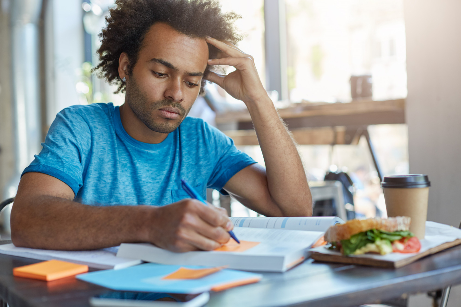 concentrated serious afro american college student with beard doing home assignment preparing for spanish lesson writing out new words from text in sticky notes during breakfast at coffee shop 2